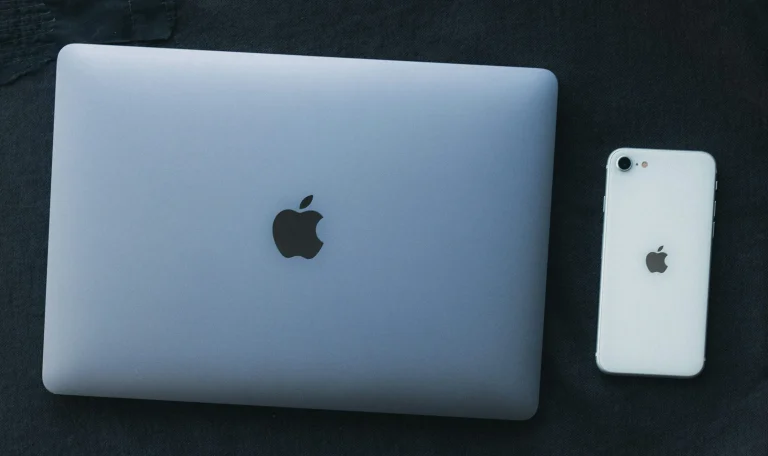 A closed silver MacBook and a white iPhone lie side by side on a dark fabric surface