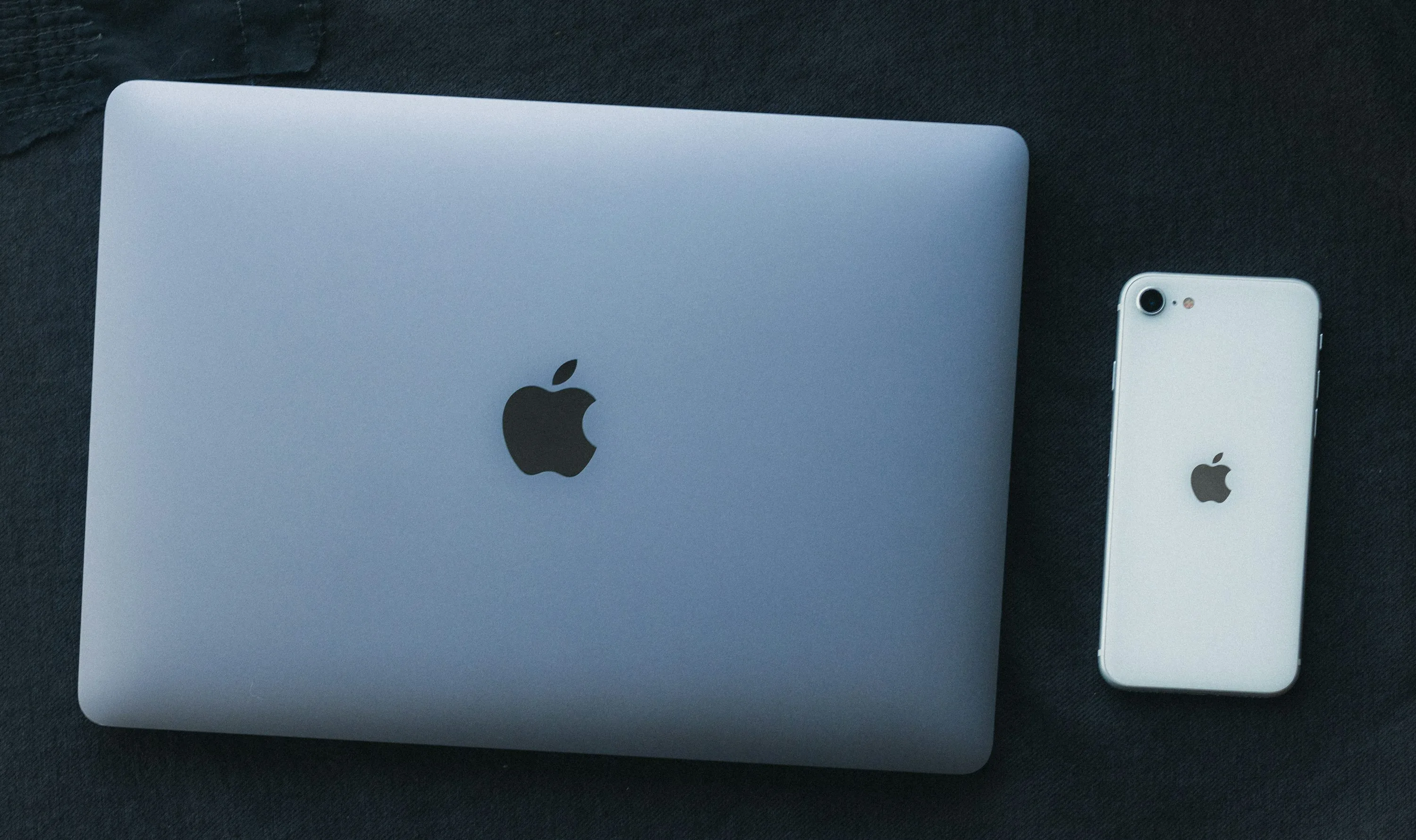 A closed silver MacBook and a white iPhone lie side by side on a dark fabric surface