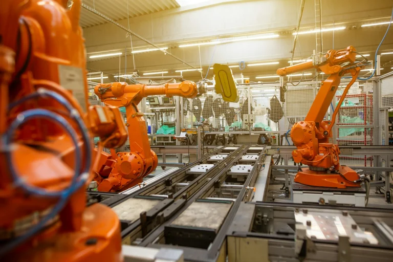 Three orange industrial robot arms stand over a factory assembly line with metal tracks and safety fencing