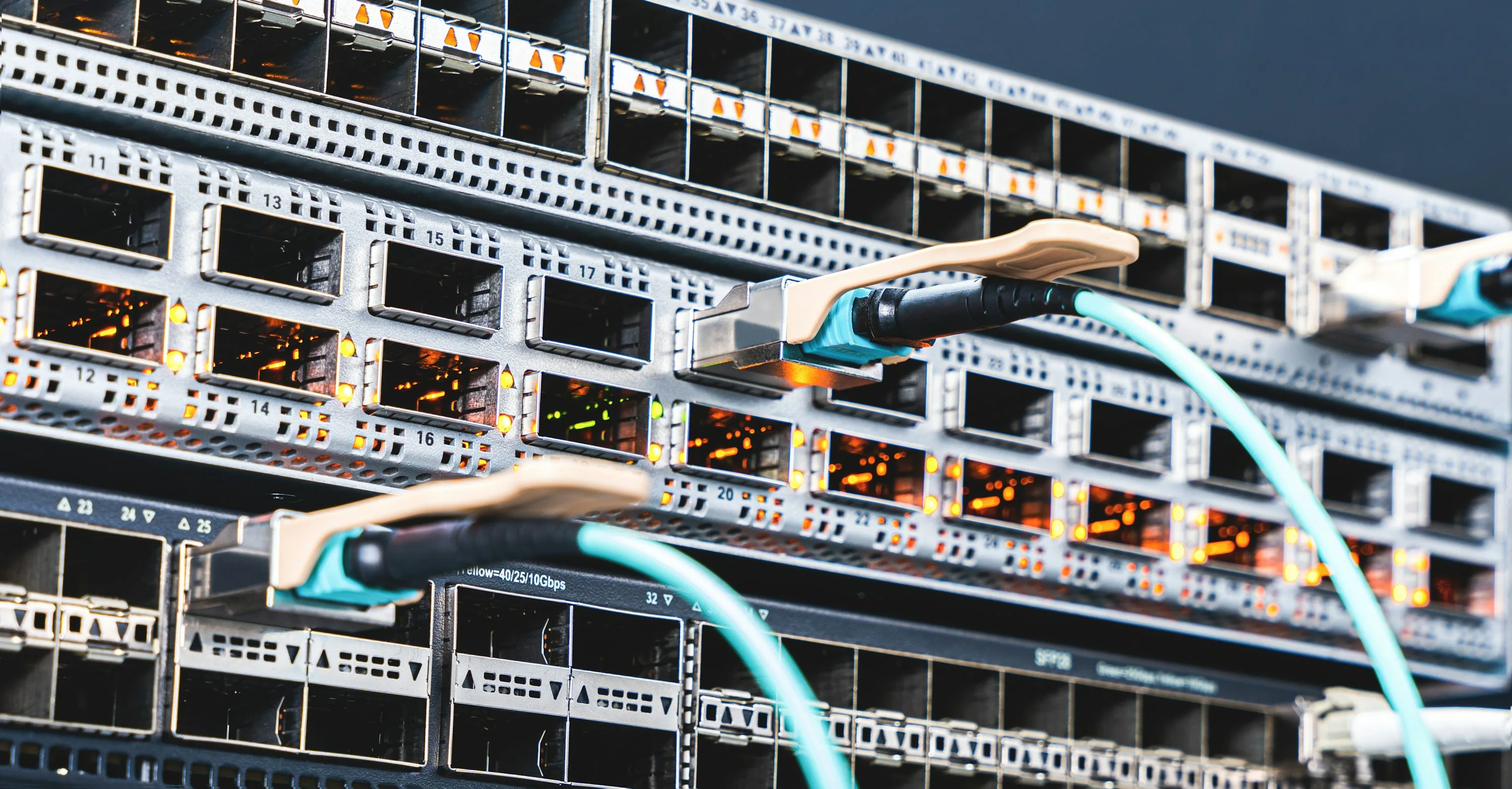 Turquoise optical cables connect to silver network switches with rows of ports and amber indicator lights