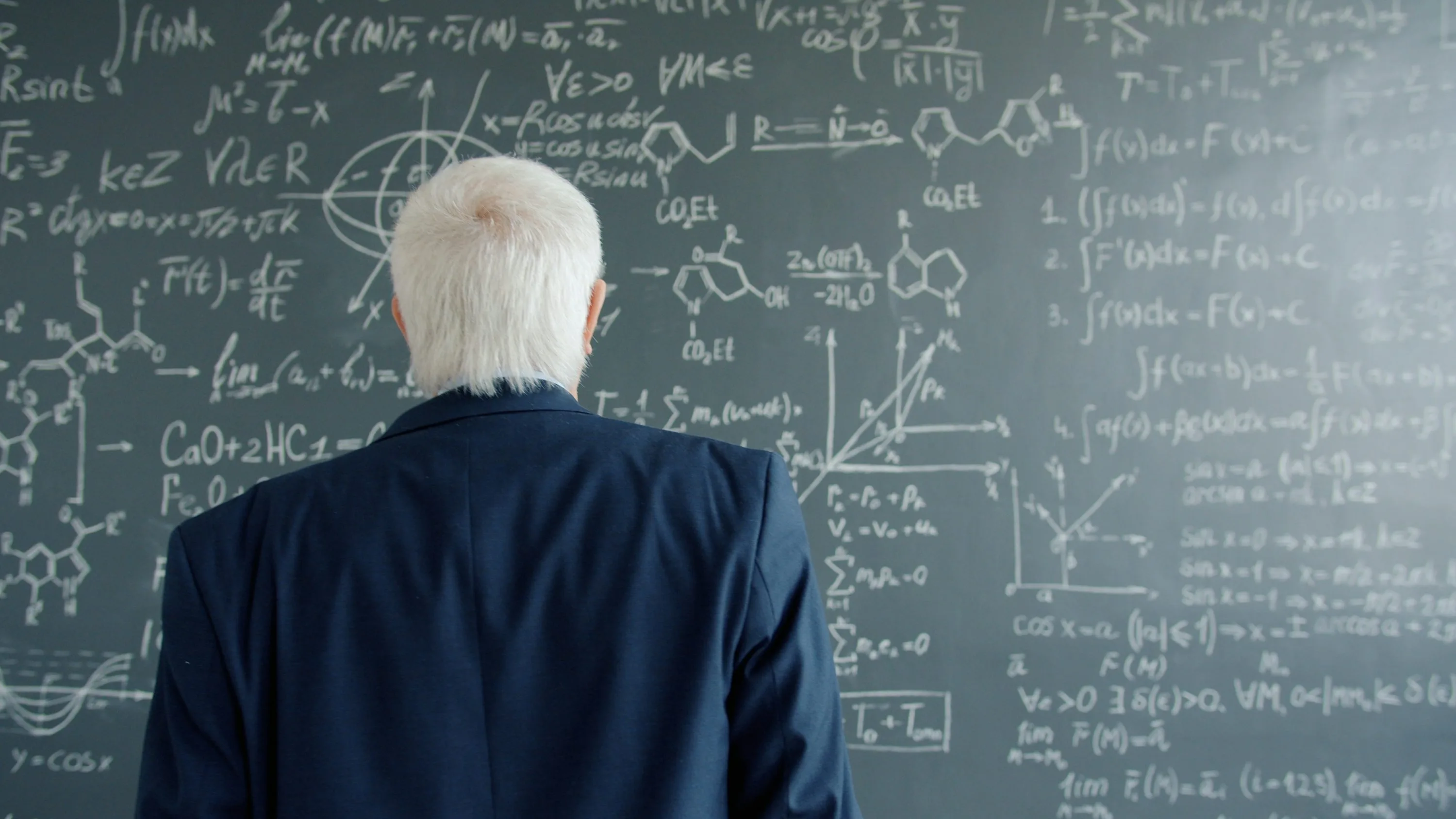 A person in a dark jacket faces a chalkboard covered with equations