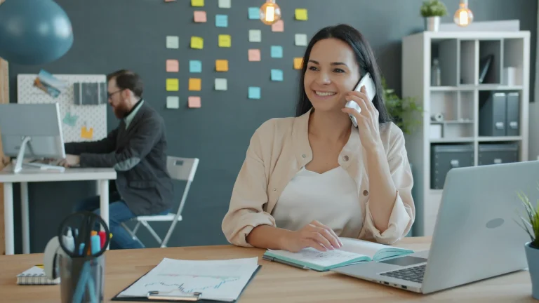 A woman holds a smartphone to her ear at a desk with an open notebook and laptop