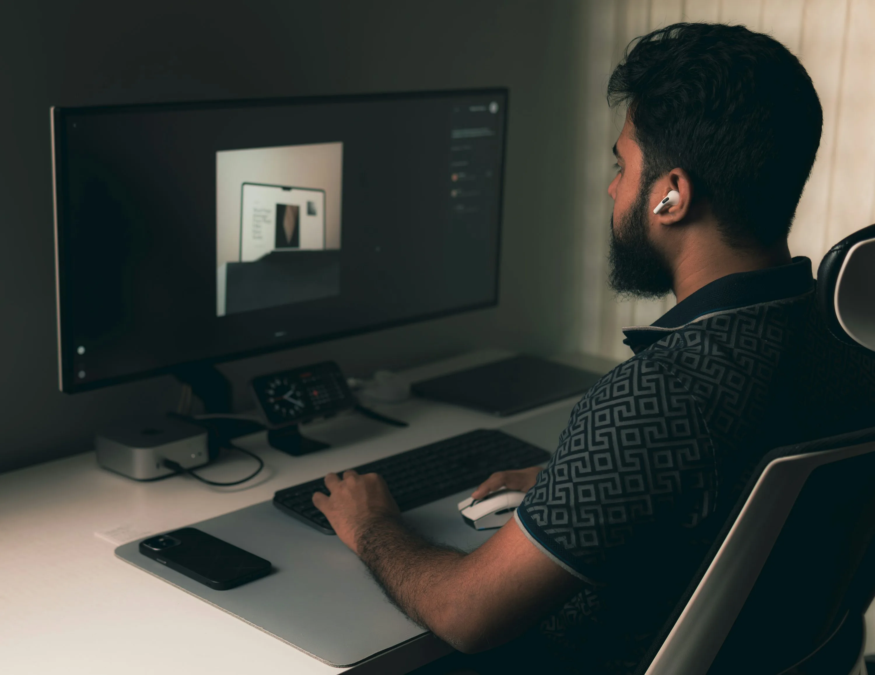 A person wearing a white earbud sits at a desk with a visual design open on a large monitor