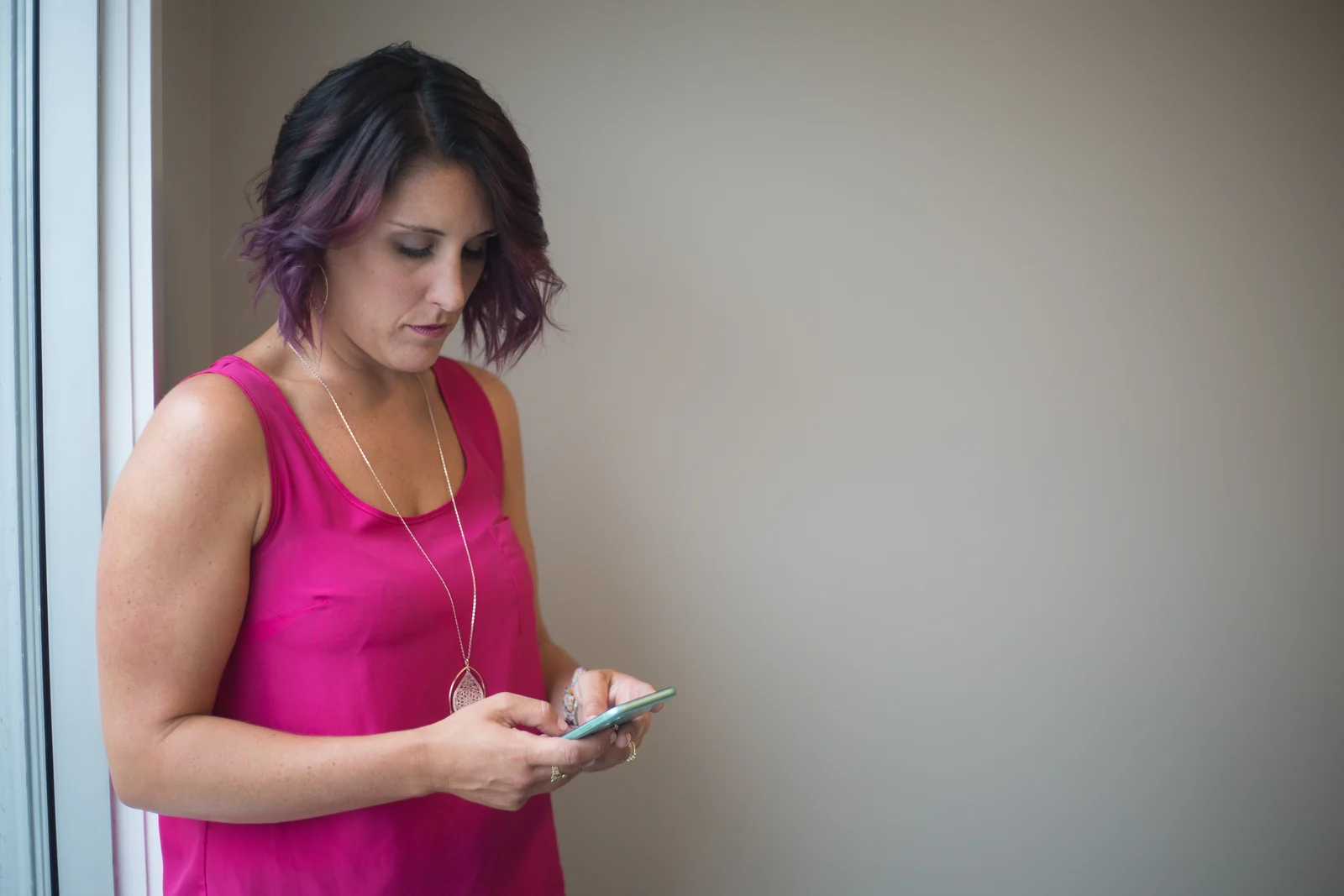 A person holds a smartphone in both hands while standing beside a window and a plain wall