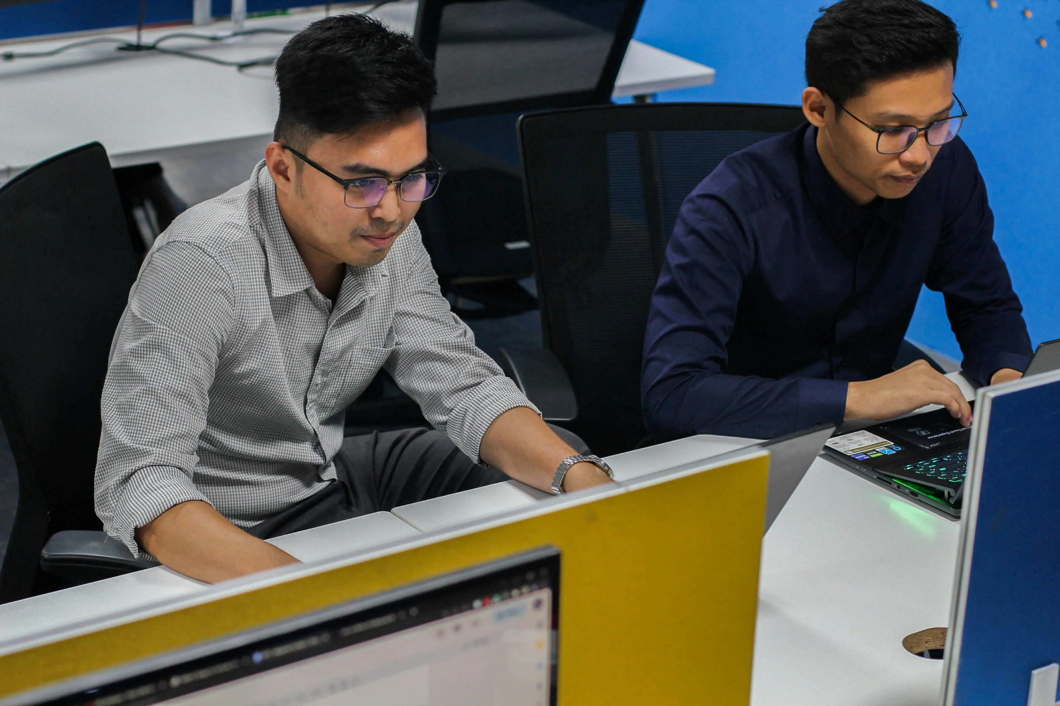 Two adults work at adjacent computers in an office with blue walls and desk partitions