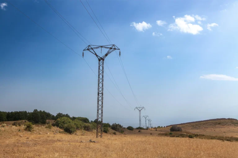 A row of metal electricity pylons carries overhead wires across dry grass beneath a blue sky