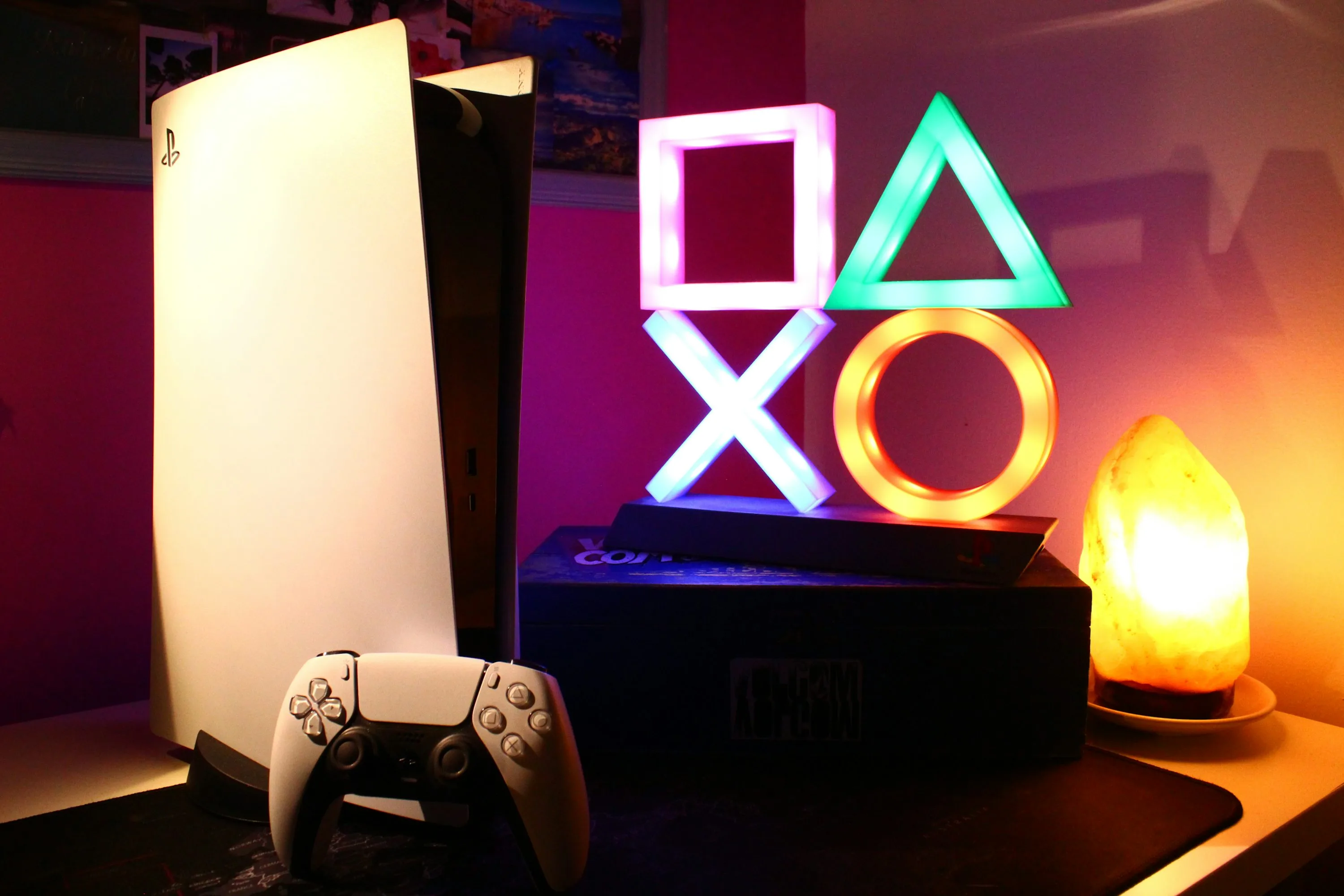 A white PlayStation 5 and controller beside colorful illuminated gaming symbols
