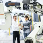 Two adults hold a robot controller between large white Kawasaki industrial robot arms in a bright robotics facility