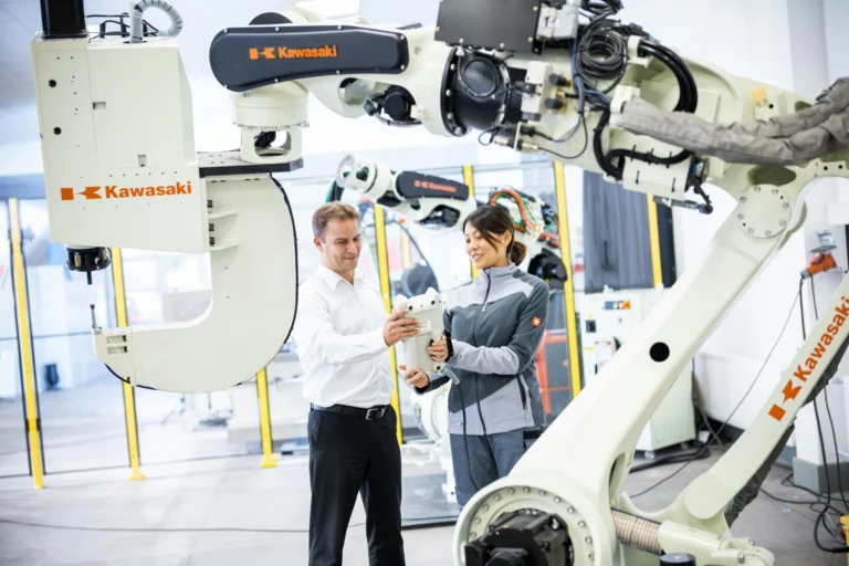 Two adults hold a robot controller between large white Kawasaki industrial robot arms in a bright robotics facility