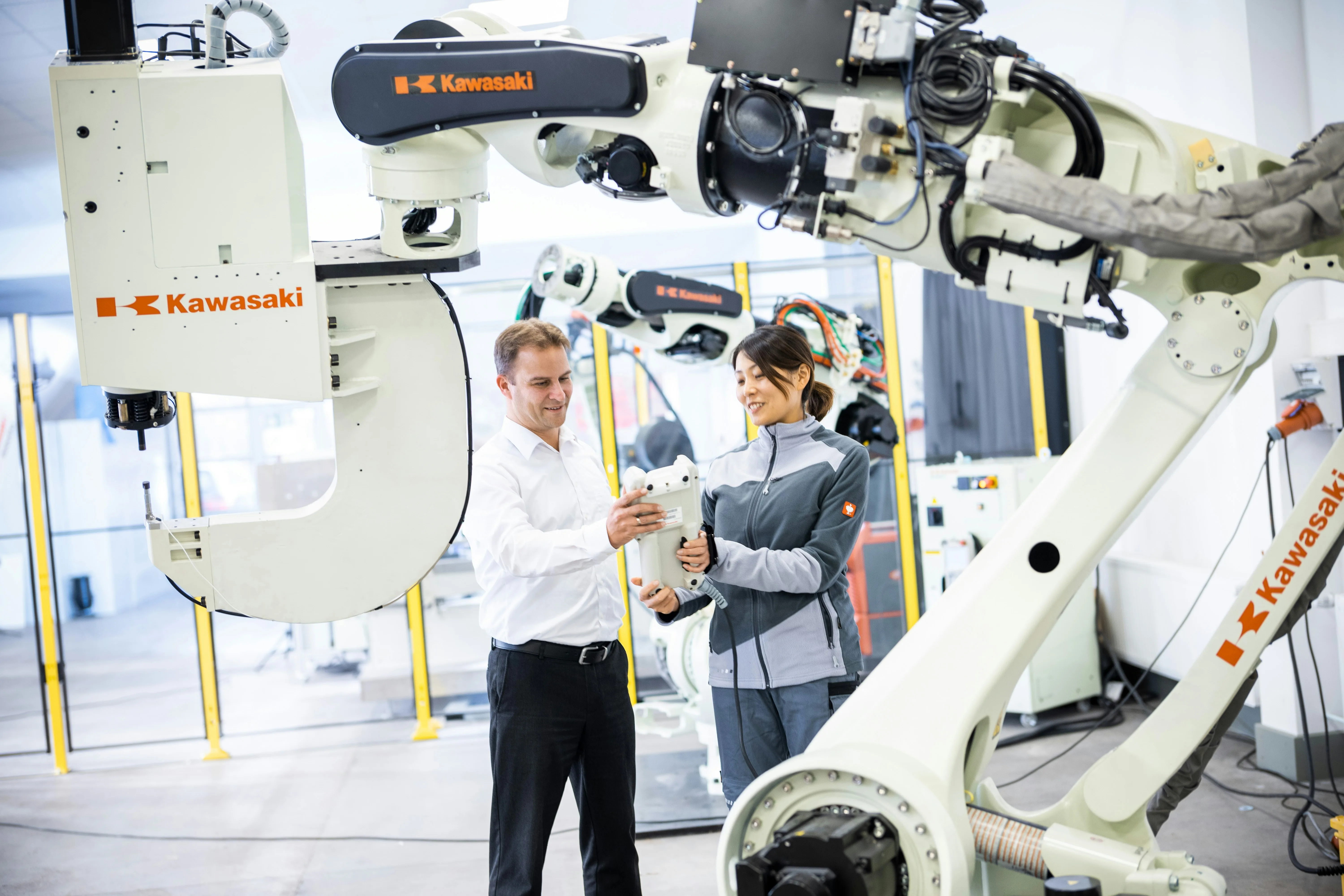 Two adults hold a robot controller between large white Kawasaki industrial robot arms in a bright robotics facility