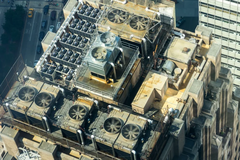 Rows of large cooling fans and pipes on an urban building rooftop