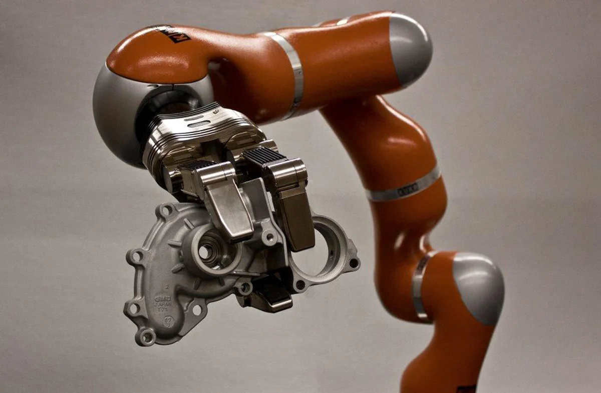 Orange robot arm with a metal gripper holding a gray mechanical component against a plain gray background