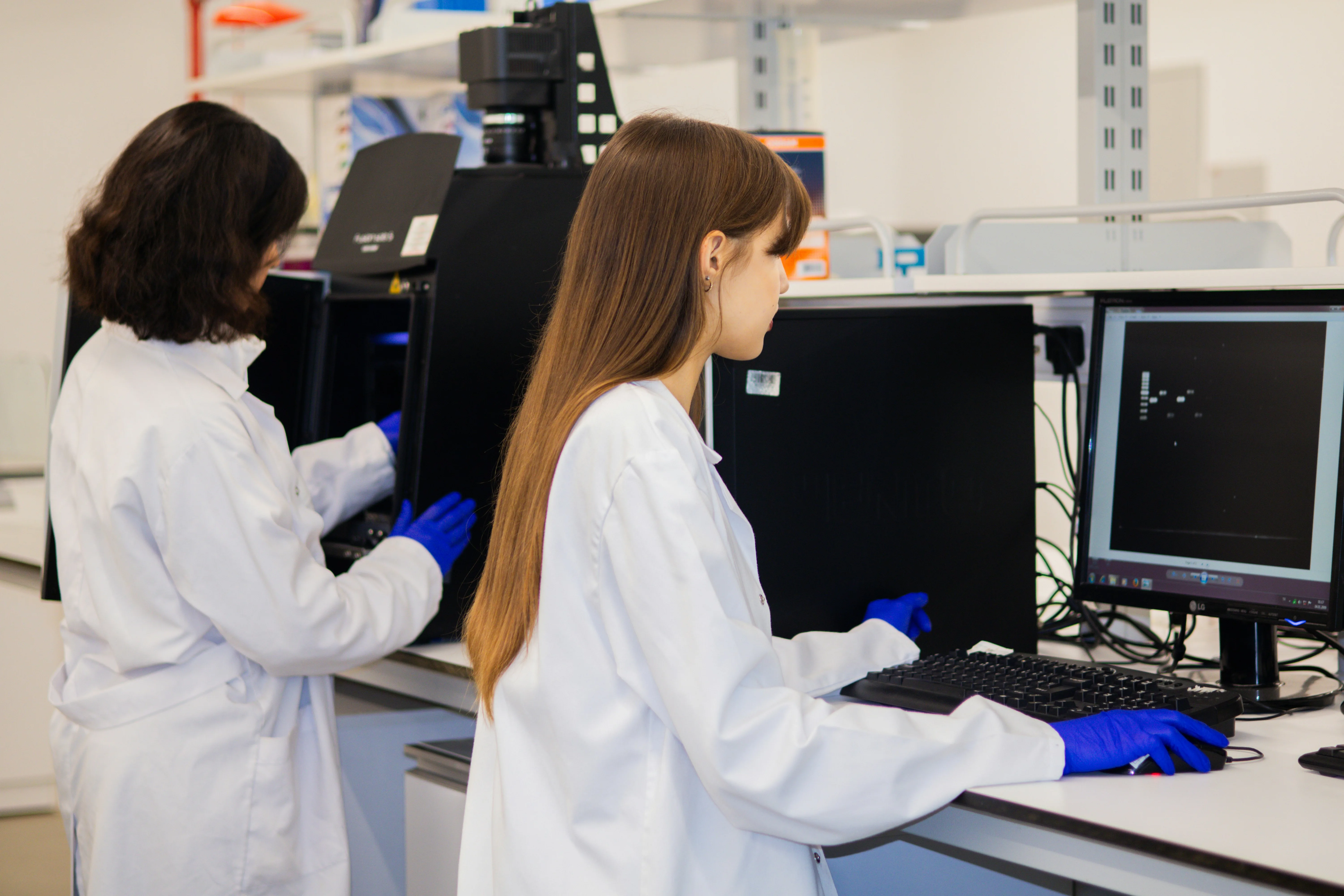 Two people in white laboratory coats and blue gloves working beside computers and scientific equipment