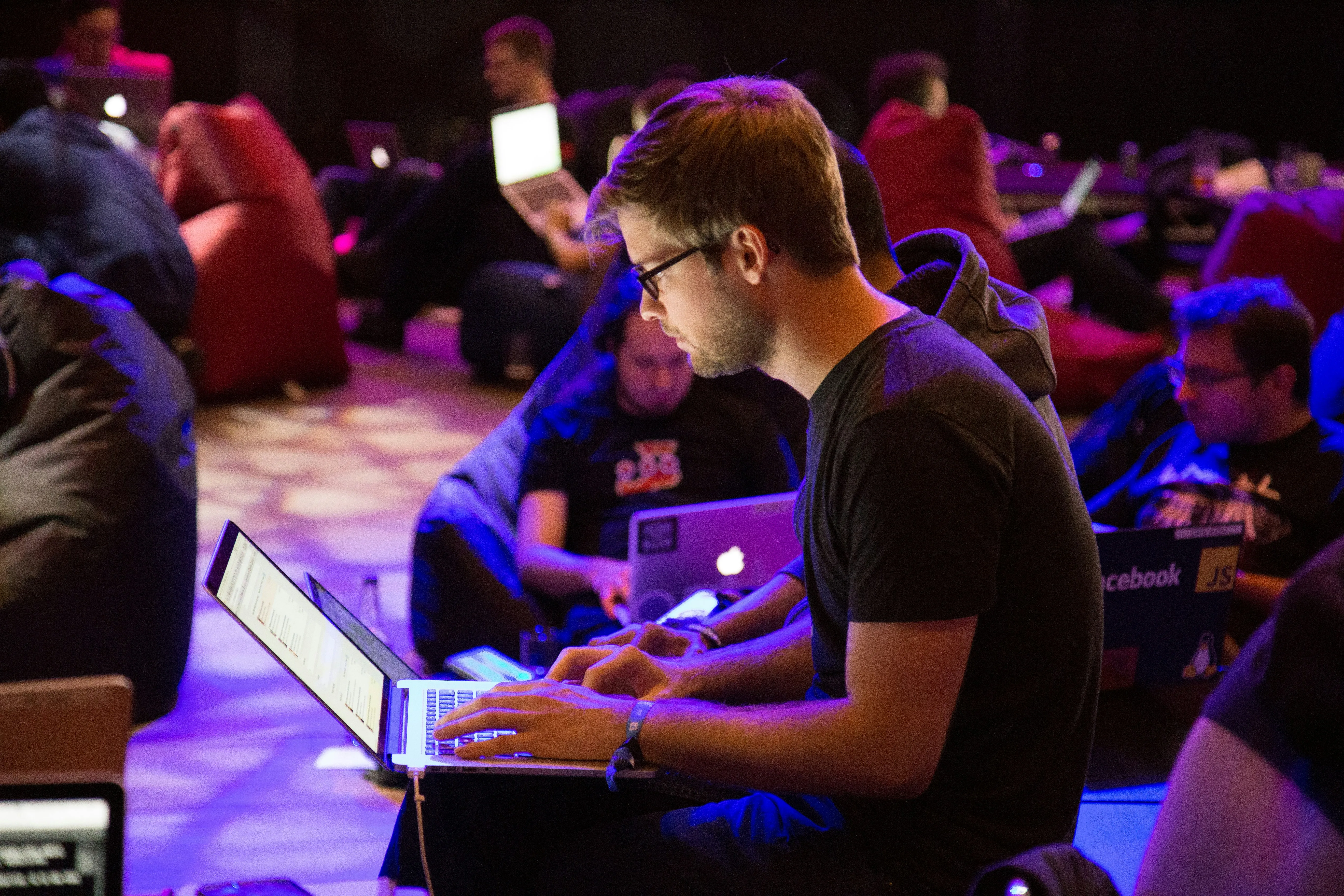 A man wearing glasses types on a laptop while other people work on computers under purple lighting