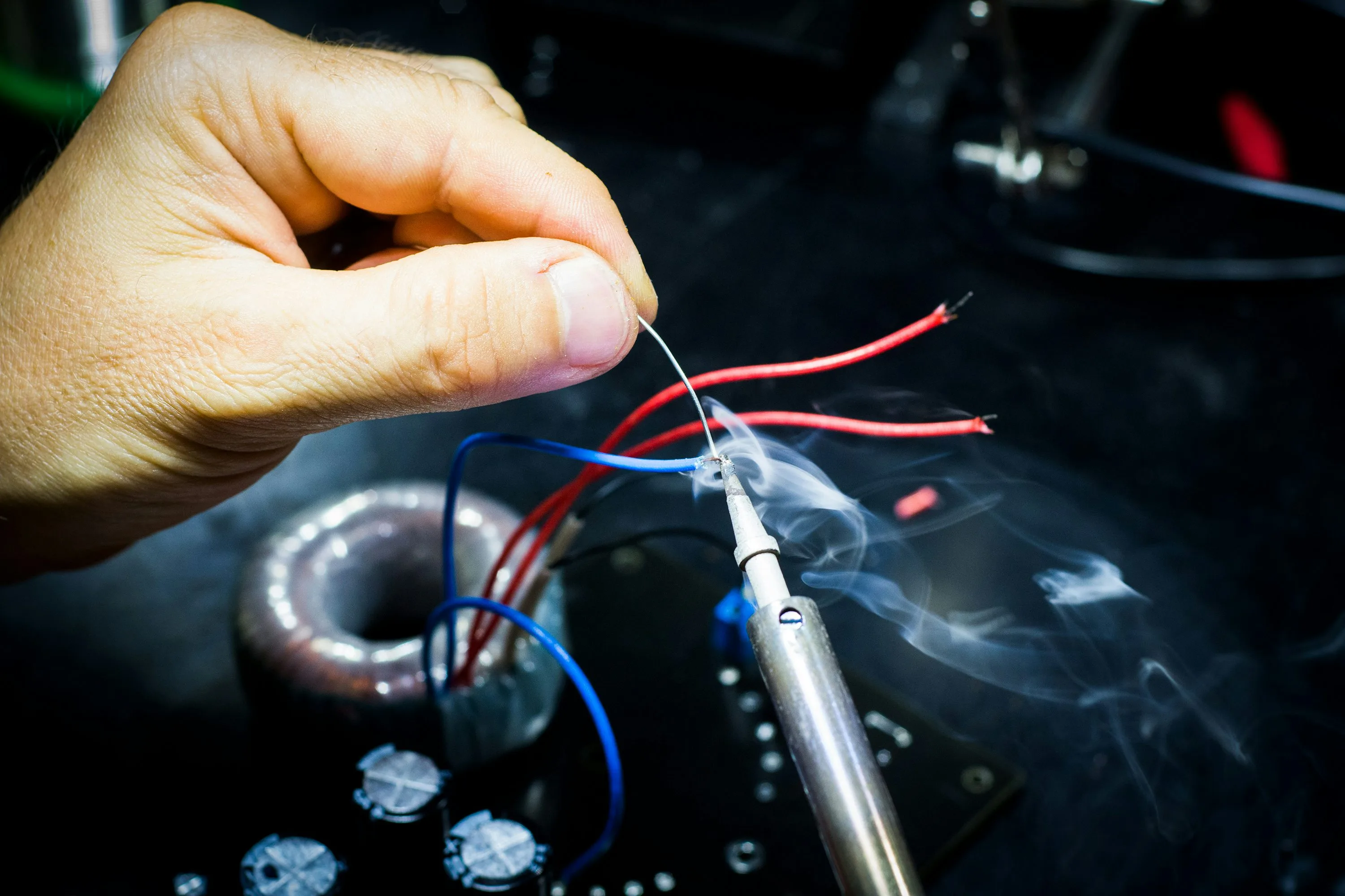 A hand feeds solder to a hot iron at a blue wire joint, with red wires and electronic components nearby