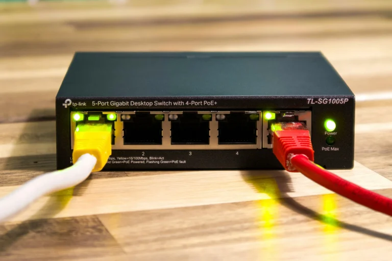 A black network switch with yellow and red Ethernet plugs and glowing green connection lights on a wooden surface