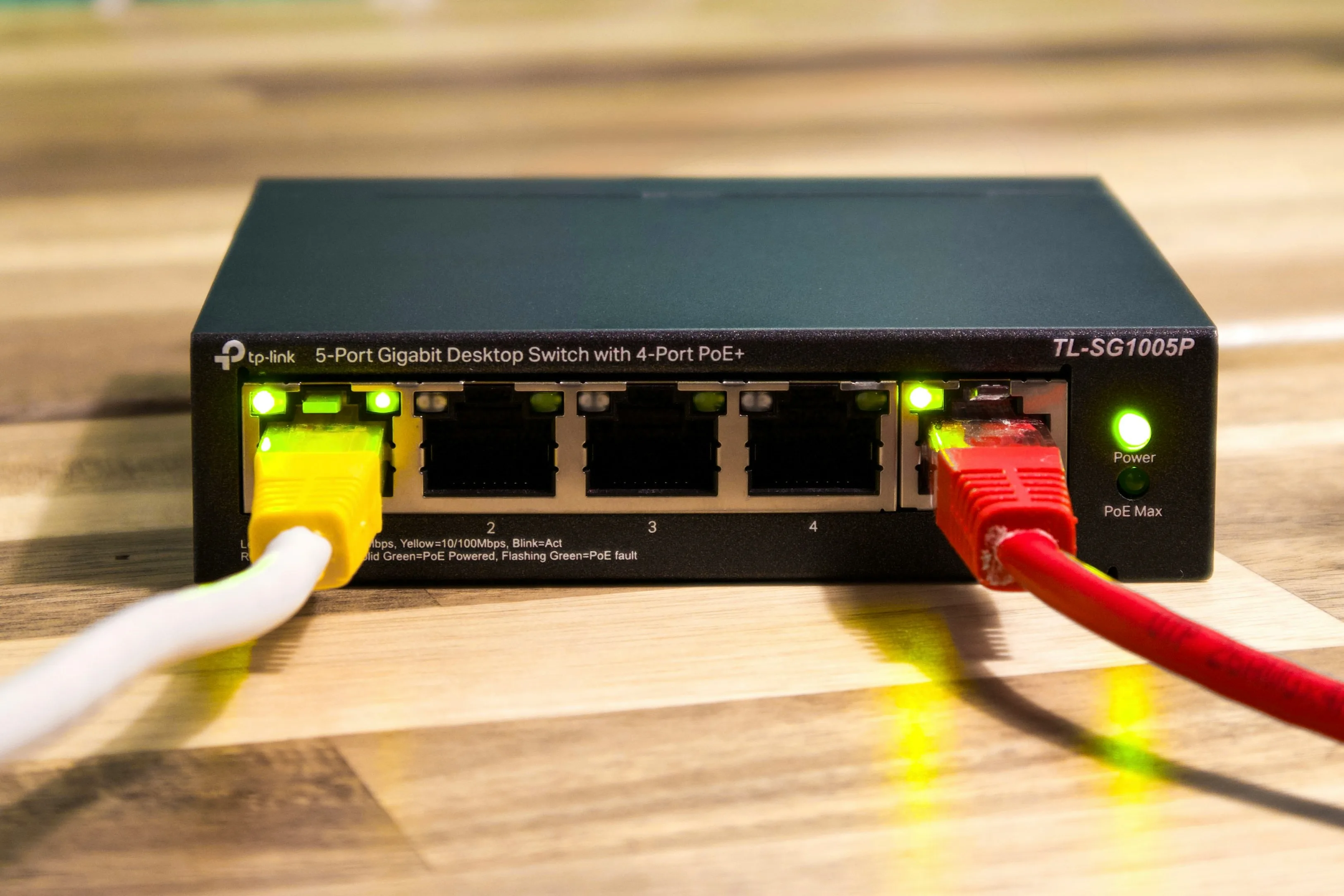 A black network switch with yellow and red Ethernet plugs and glowing green connection lights on a wooden surface