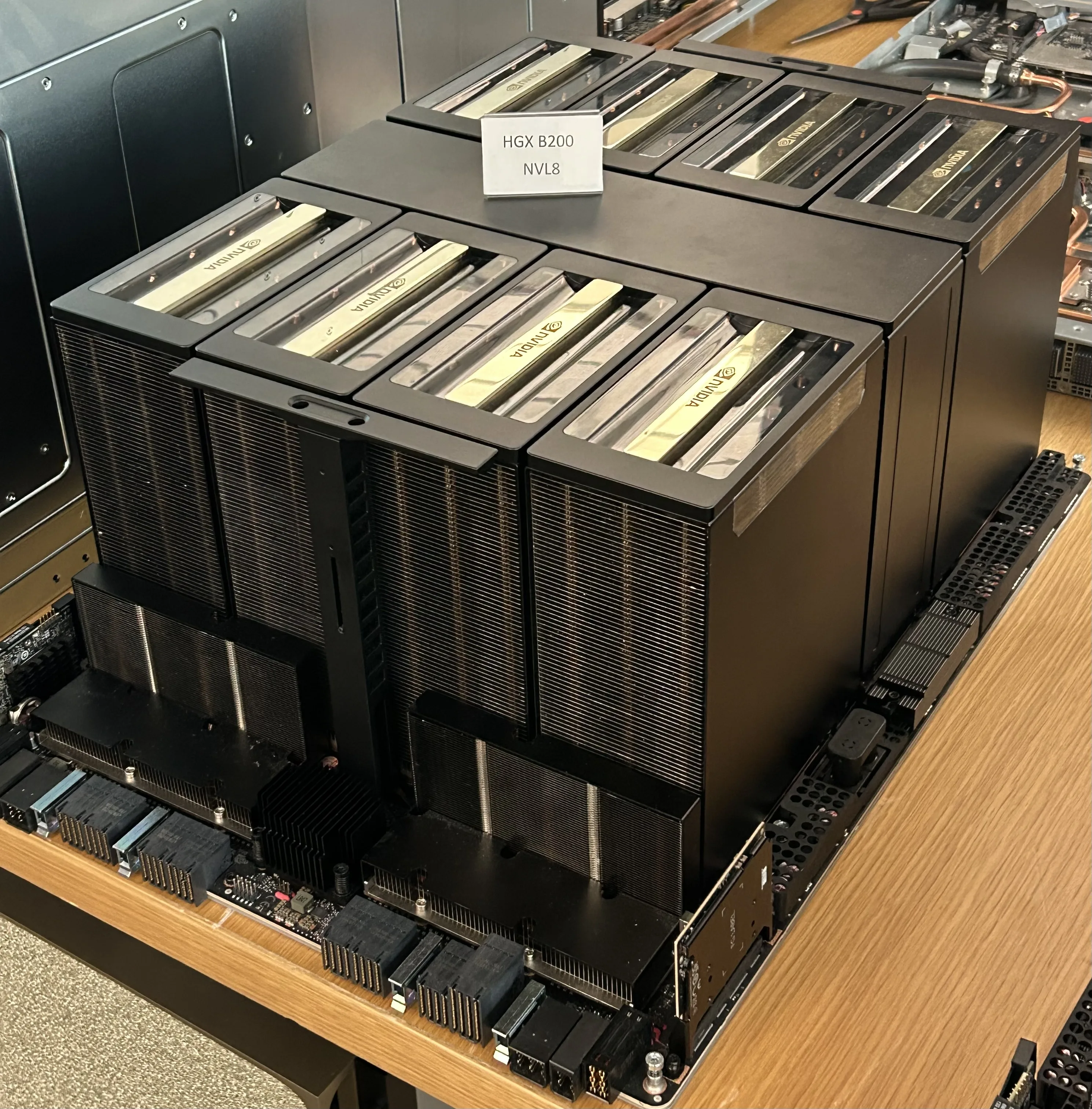 Eight black GPU cooling assemblies on an NVIDIA HGX B200 board displayed on a wooden table