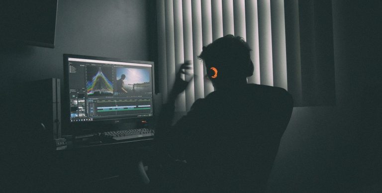 A person in silhouette wearing headphones editing video on a computer in a dark room