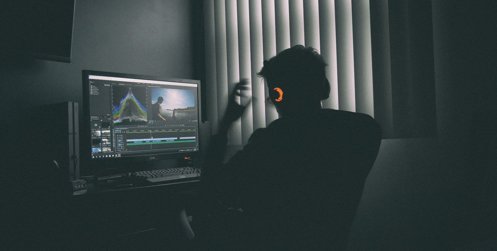 A person in silhouette wearing headphones editing video on a computer in a dark room