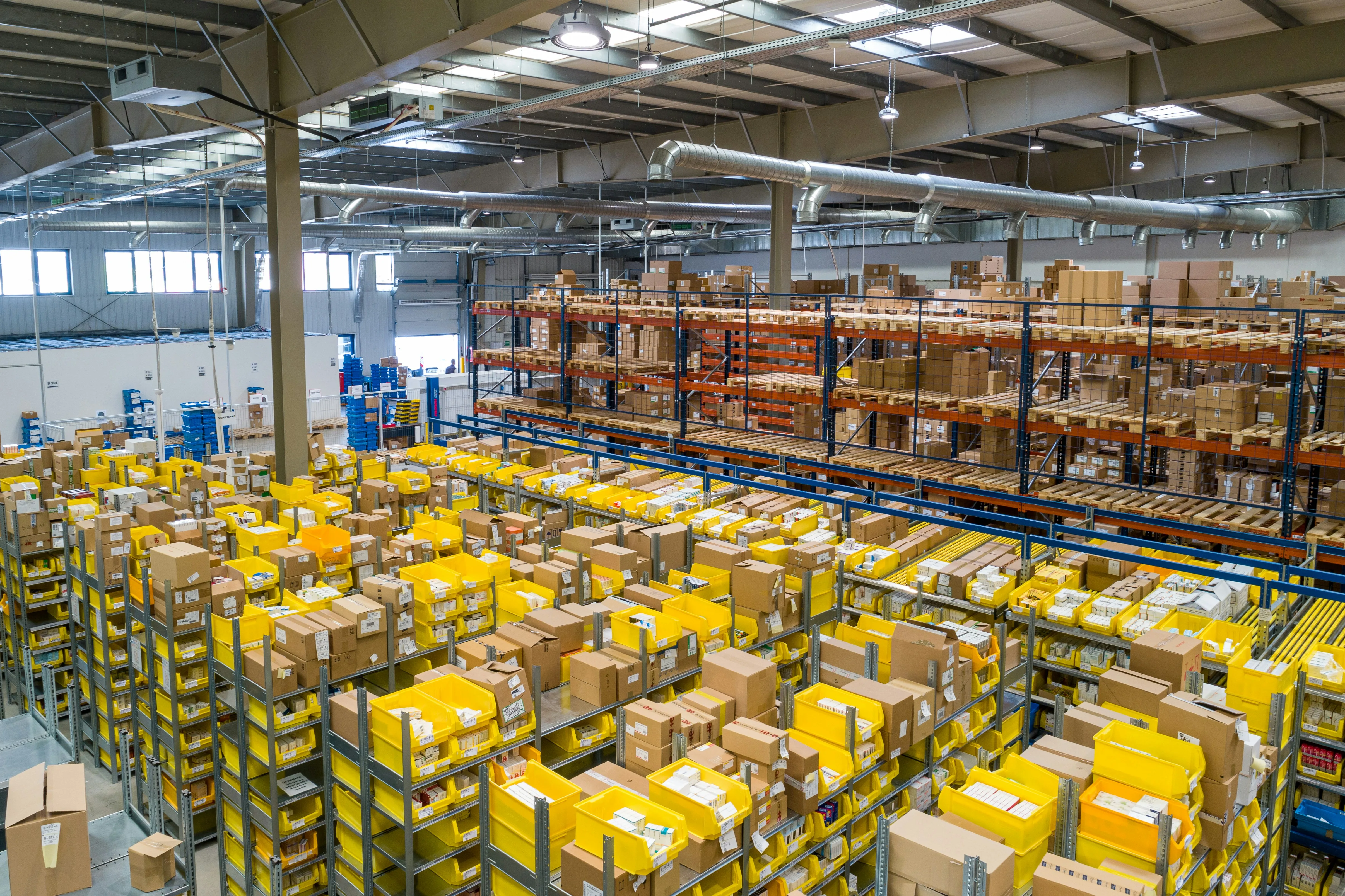 Yellow storage bins and cardboard boxes fill shelves beneath the roof of a warehouse