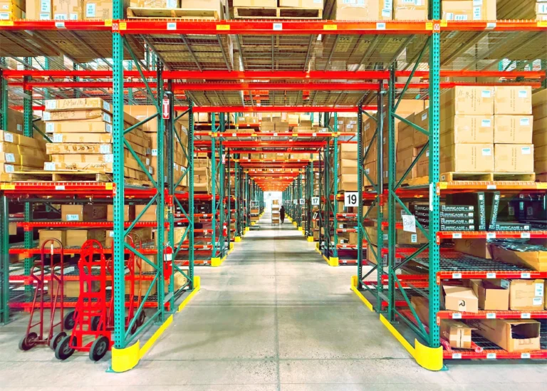 A warehouse aisle runs between red and green shelving loaded with boxes, with hand trucks in the foreground and a distant worker