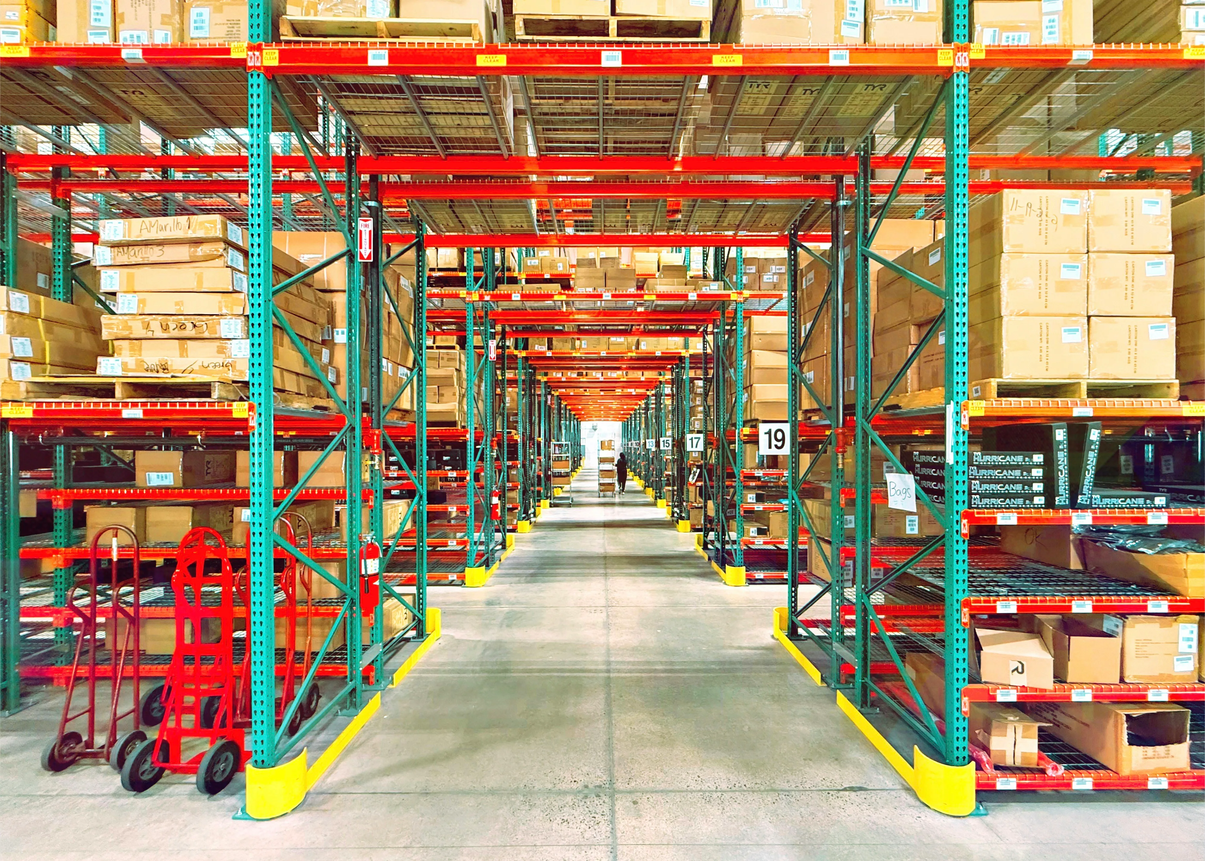 A warehouse aisle runs between red and green shelving loaded with boxes, with hand trucks in the foreground and a distant worker